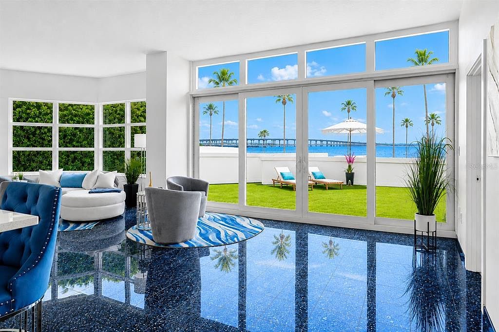The main living room of Residence 109 with a wall of windows framing Sarasota Bay and the fishing pier, curved seating, and a polished Blue Pearl granite floor reflecting the room