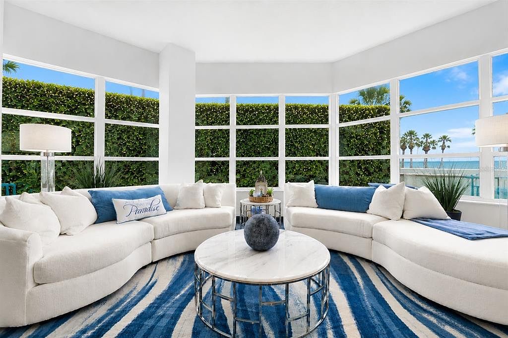 A living area with white curved sectionals around a marble coffee table, a blue and white rug, a Paradise throw pillow, and a tall green hedge with the bay visible through the windows