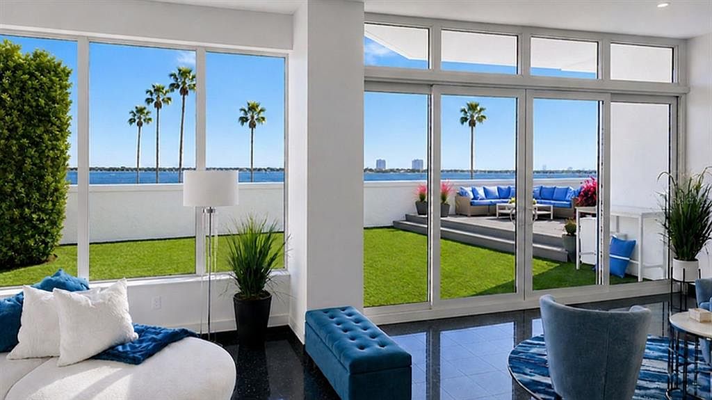A sitting area opening through full-height sliding glass doors to the terrace lawn and a blue outdoor sofa, with palm trees and Sarasota Bay beyond