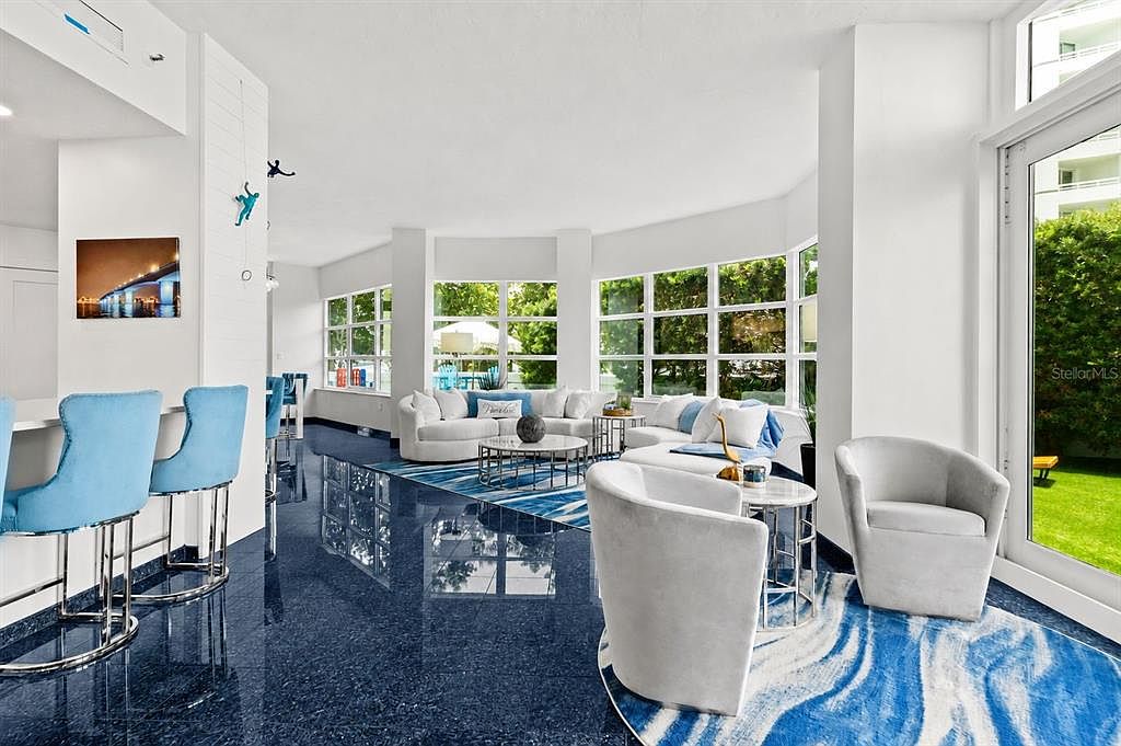 A wide great room with light-blue bar stools at a counter, curved white sofas, a framed artwork of the Ringling Bridge at night, and a Blue Pearl granite floor