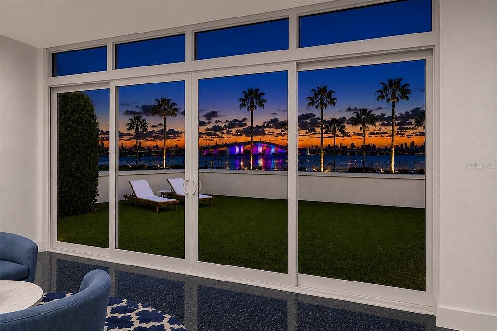 Interior at twilight: a wall of sliding glass doors framing the Ringling Bridge lit in purple at sunset over Sarasota Bay, with two chaise loungers on the terrace lawn outside