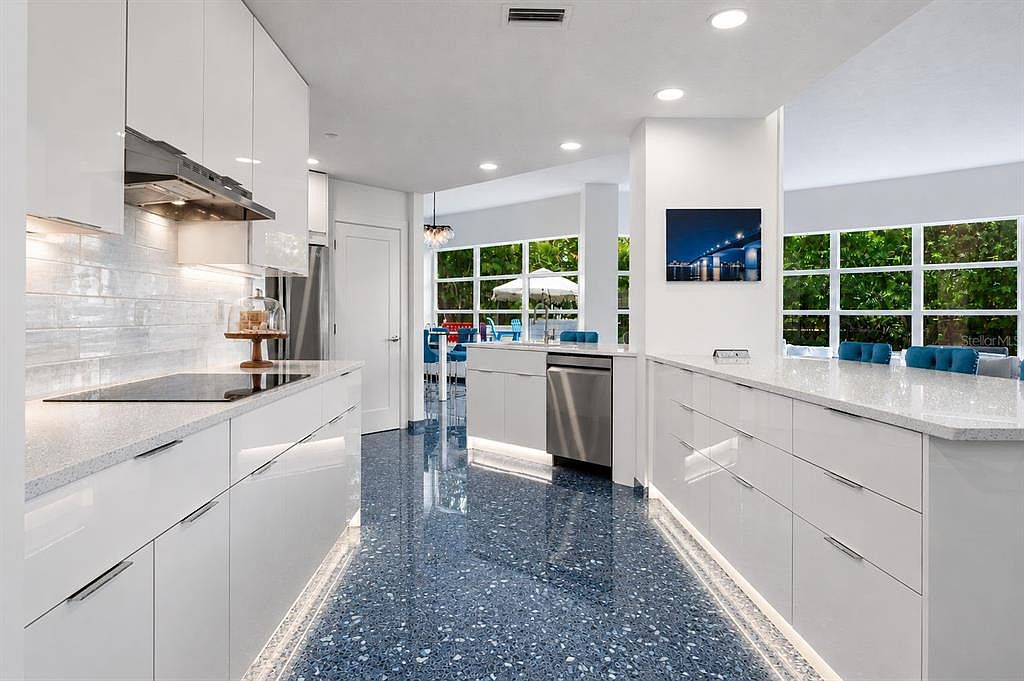 The waterfront main kitchen with white flat-panel cabinets, light quartz counters, stainless appliances, a Blue Pearl granite floor, and a dining area with blue stools and windows to the garden beyond