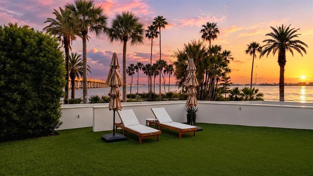 The private bayfront terrace at sunset, a wide green lawn with teak chaise loungers, palm trees, Sarasota Bay, and the Ringling Bridge under a vivid pink and orange sky